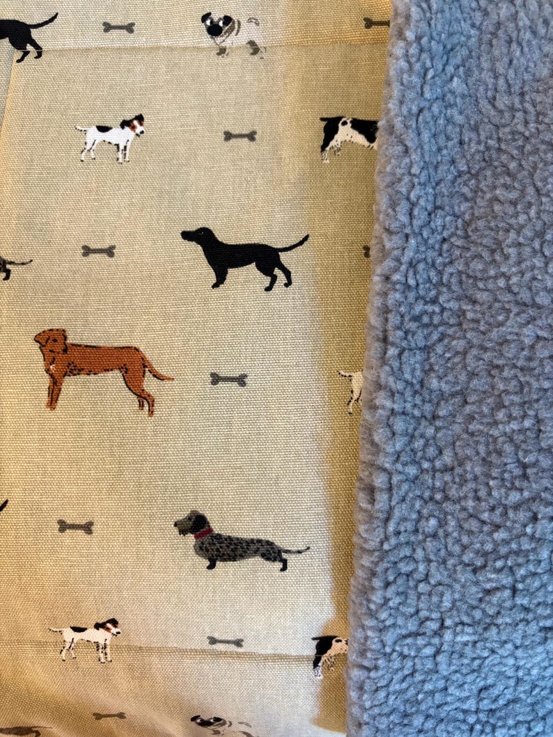 May include: A beige fabric with a repeating pattern of various dog breeds and bone shapes. The fabric is next to a fluffy, light blue material. The dogs are in black, brown, and white. The overall theme is pet-related.