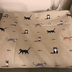 May include: A beige fabric with a repeating pattern of black, white, and gray cats. The cats are in various poses, including sitting, standing, and walking. There are also small brown fishbone shapes scattered throughout the fabric.