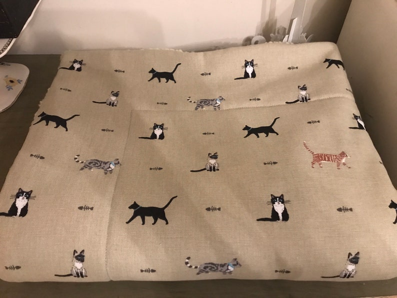 May include: A beige fabric with a repeating pattern of black, white, and gray cats. The cats are in various poses, including sitting, standing, and walking. There are also small brown fishbone shapes scattered throughout the fabric.