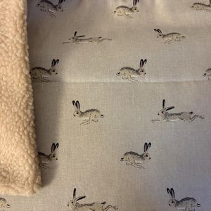 Puede incluir: Tela azul claro con un patrón repetido de liebres corriendo en gris y negro. En el lado izquierdo se ve una tela cremosa y esponjosa. El diseño es adecuado para la decoración del hogar o proyectos textiles.