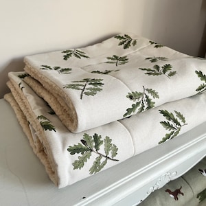 Puede incluir: Dos mantas plegadas, de color crema, con un estampado de hojas de roble verdes. Las mantas tienen un reverso suave, de color crema, similar a la sherpa. Las mantas están apiladas sobre una cómoda blanca ornamentada.