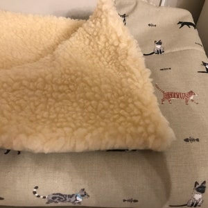 May include: A beige cat bed with a removable, fluffy cream-colored fleece liner. The bed has a green fabric exterior with a repeating pattern of black, white, and brown cats and fish bones.