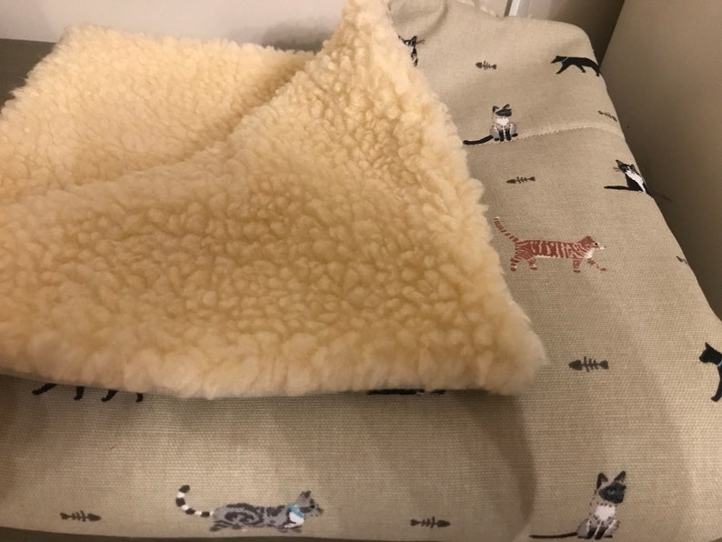 May include: A beige cat bed with a removable, fluffy cream-colored fleece liner. The bed has a green fabric exterior with a repeating pattern of black, white, and brown cats and fish bones.