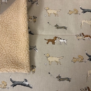 Puede incluir: Una manta con temática de perros con un forro de forro polar sherpa beige claro. La tela superior es de color gris claro con varias razas de perros en beige, marrón y gris, jugando con pelotas. La manta está doblada, mostrando el suave forro polar texturizado.