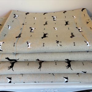 May include: Stacked dog beds with a light tan background and a repeating pattern of black and white dogs and small bone shapes. The beds are rectangular and appear to be made of a soft fabric.