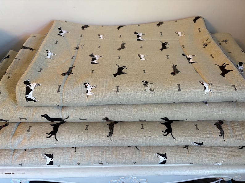May include: Stacked dog beds with a light tan background and a repeating pattern of black and white dogs and small bone shapes. The beds are rectangular and appear to be made of a soft fabric.