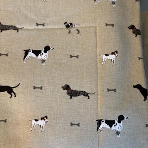 May include: Tan fabric with a repeating pattern of various dog breeds and small gray bone shapes. The dogs are in black, white, and brown. The fabric is folded over, showing the pattern on the front and the side.