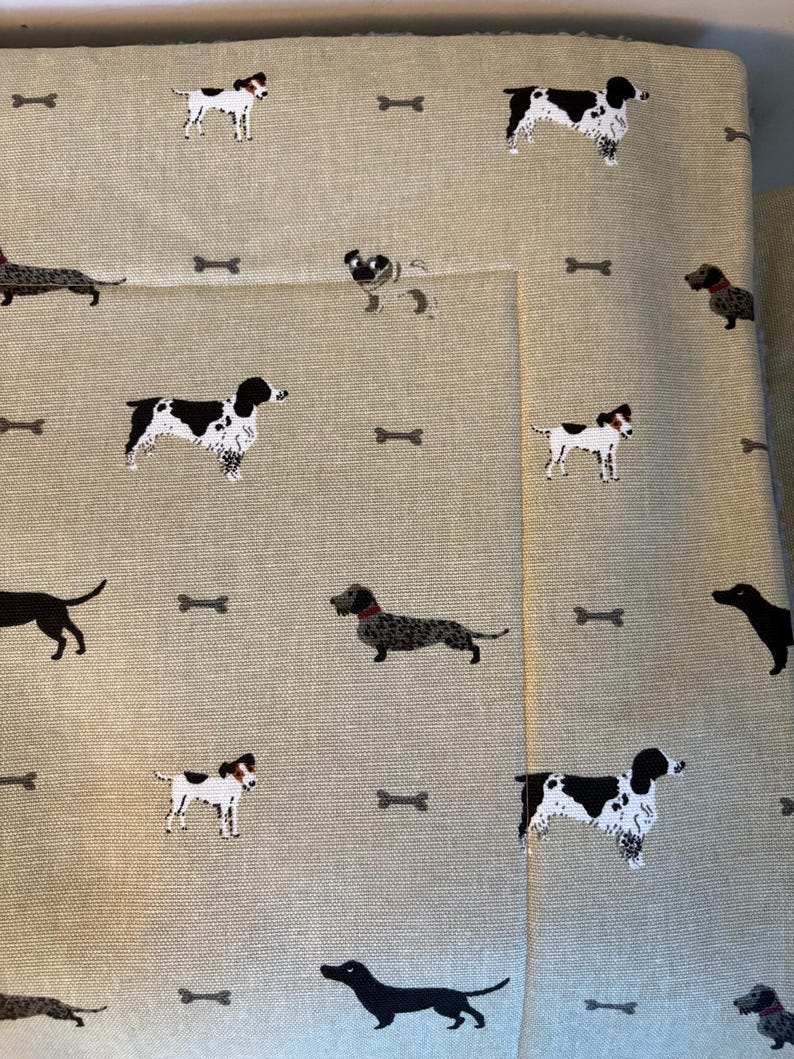 May include: Tan fabric with a repeating pattern of various dog breeds and small gray bone shapes. The dogs are in black, white, and brown. The fabric is folded over, showing the pattern on the front and the side.
