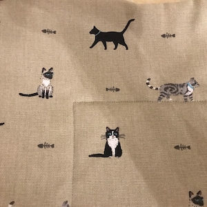 May include: A beige fabric cushion with a repeating pattern of black and white cats and fish bones. The cats are in various poses, including sitting, standing, and walking.