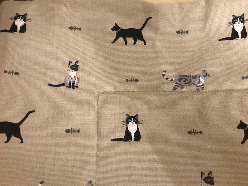 May include: A beige fabric cushion with a repeating pattern of black and white cats and fish bones. The cats are in various poses, including sitting, standing, and walking.