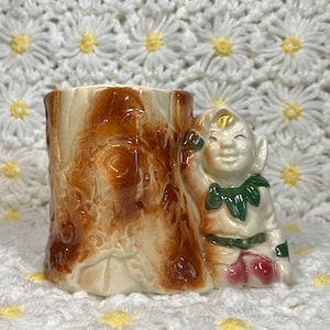 Op de afbeelding: Een keramische plantenpot in de vorm van een boomstronk met een glimlachende kabouter die erachter vandaan kijkt. De kabouter draagt ​​een groene hoed en een rood-groene outfit.