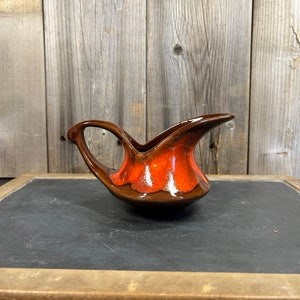 Puede incluir: Una jarra de crema o salsa de cerámica marrón y naranja con un asa curva única.