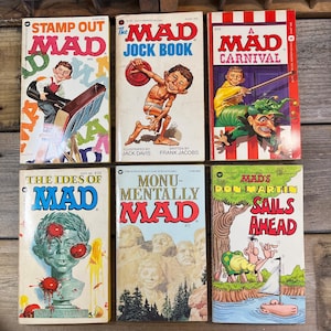 Könnte beinhalten: Eine Sammlung von sechs Vintage-Comic-Heften des Mad Magazine. Die Cover zeigen farbenfrohe Illustrationen und fetten Text, darunter Titel wie "Stamp Out Mad", "The Mad Jock Book" und "A Carnival". Die Bücher sind auf einer Holzoberfläche angeordnet.