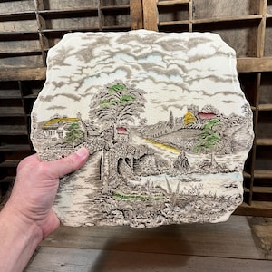 Può includere: Un piatto da portata in ceramica vintage con un design in rilievo di una scena di campagna. La scena presenta un ponte di pietra, un sentiero tortuoso, un piccolo villaggio e una carrozza trainata da cavalli. Il piatto è decorato con colori marrone, verde e giallo.