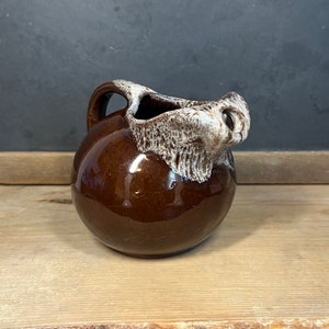 May include: A small, round, brown ceramic jug with a handle. The top edge and spout are a lighter, textured colour. The jug sits on a wooden surface.