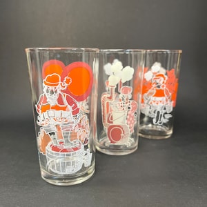 May include: Three clear glass drinking glasses with red and white illustrations of Santa Claus, a snowman, and a person in a red and white suit. The glasses are on a black surface.