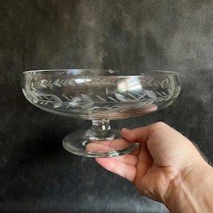 Peut inclure: Un bol en verre transparent sur un piédestal avec un motif de feuilles gravé délicatement. Le bol est sur un piédestal rond et court.