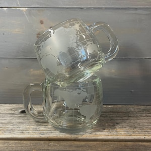 Puede incluir: Dos tazas de vidrio transparente con asas están apiladas. Cada taza presenta un diseño de mapa del mundo grabado en la superficie, con líneas de cuadrícula que indican latitud y longitud. Las tazas son transparentes.
