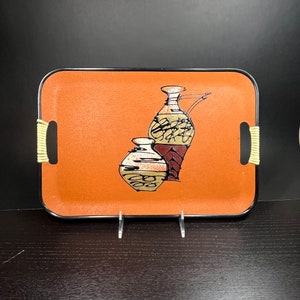 Puede incluir: Una bandeja rectangular naranja con ribete negro y dos asas beige. La bandeja presenta un diseño negro y dorado de dos macetas de cerámica estilizadas.
