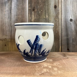 May include: A white ceramic onion holder with blue and white Delftware designs of a windmill and houses. The holder has several holes for ventilation.