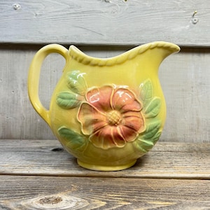 Puede incluir: Una jarra de cerámica amarilla con un diseño floral. La jarra presenta una gran flor en relieve en tonos naranja y rosa, rodeada de hojas verdes. El asa y el borde también son amarillos.