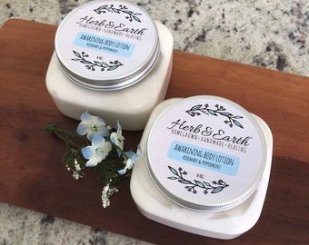 Pode incluir: Dois frascos brancos de loção corporal com tampas prateadas. Os frascos são etiquetados com "Herb & Earth" e "Awakening Body Lotion Rosemary & Peppermint" com um design floral. Os frascos estão sobre uma superfície de madeira.