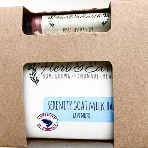 May include: A brown cardboard box with a cut-out window showing a bar of lavender goat milk soap and a tube of rose chapstick. The soap is labeled "Serenity Goat Milk Bar Lavender" and is certified as a South Carolina product. The chapstick is labeled "Herb & Earth Rose Chapstick + SPF".