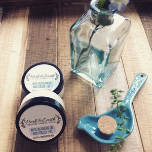 May include: Two jars of Herb & Earth anti-aging facial moisturizer with SPF. The jars are on a wooden surface with a blue bird-shaped dish and a clear glass bottle with a flower.