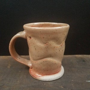 May include: A brown ceramic mug with a textured, uneven surface and a white base. The mug has a handle and is slightly wider at the top than the bottom.