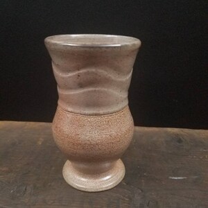 May include: A light brown ceramic vase with a textured surface and a wavy design around the middle.