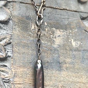 May include: A silver chain necklace with a gold clasp and a long, dark grey baroque pearl pendant.