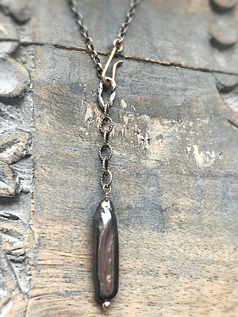 May include: A silver chain necklace with a gold clasp and a long, dark grey baroque pearl pendant.