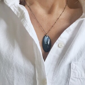 Peut inclure: Un collier en cha&icirc;ne argent avec un pendentif ovale bleu fonc&eacute;. Le pendentif a un motif stri&eacute;.