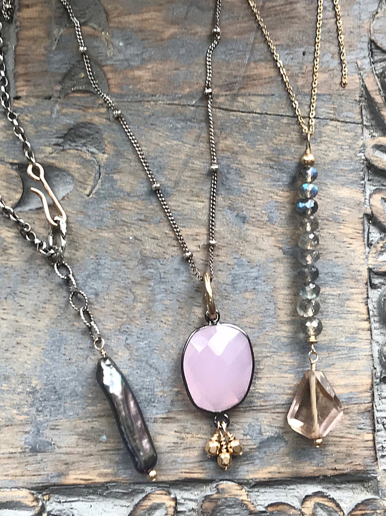 May include: Three necklaces with different pendants. The first necklace has a dark grey pearl pendant. The second necklace has a pink gemstone pendant with gold accents. The third necklace has a clear quartz pendant with a grey crystal bead chain.