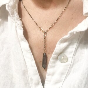 May include: A silver chain necklace with a long, dark grey pendant hanging from it. The pendant is a smooth, rectangular shape.