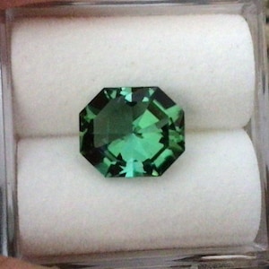 May include: A green gemstone with a faceted octagonal shape, displayed in a white box.