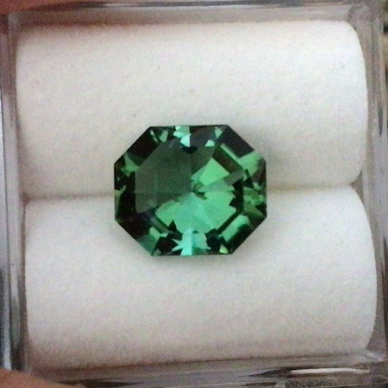 May include: A green gemstone with a faceted octagonal shape, displayed in a white box.