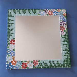 Könnte beinhalten: Ein rechteckiger Spiegel mit einem Mosaikrahmen. Der Rahmen besteht aus kleinen, bunten Fliesen in den Farben Grün, Blau, Rot, Orange und Weiß. Die Fliesen sind in einem floralen Muster angeordnet.