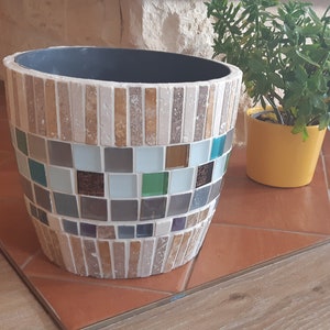 May include: A round, decorative planter made of mosaic tiles. The planter is made of small, square tiles in shades of brown, beige, blue, green, and white. The planter is sitting on a tiled floor.