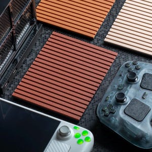 May include: A black gaming console, a handheld gaming device with green buttons, a translucent gray game controller, and three rectangular panels with horizontal slats in shades of light beige, orange, and dark reddish-brown.