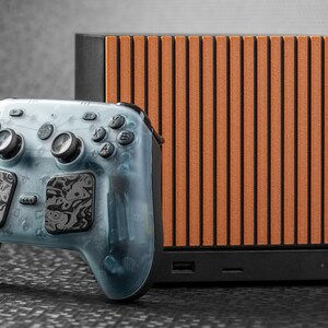 May include: A rectangular gaming console with a black base and a front panel featuring vertical orange slats, paired with a translucent light blue wireless game controller that has black and white marbled touchpads and standard buttons.