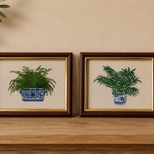 May include: Two rectangular framed embroideries featuring green potted ferns. The left piece shows a dense fern in a rectangular blue and white patterned planter. The right piece shows a palm-like fern in a round blue and white patterned pot.