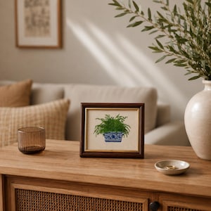 Small framed fern embroidery artwork displayed on a wooden console table in a cozy living room, styled with neutral decor and natural greenery.