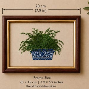 May include: A framed embroidery featuring a green fern in a blue and white patterned rectangular planter. The dark brown wooden frame has a gold inner border. The overall dimensions of the frame are 7.9 inches by 5.9 inches as indicated.