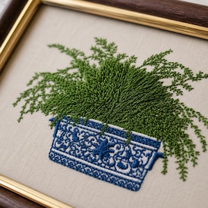 Close-up of intricate hand embroidery stitches showing a detailed green fern and ornate blue chinoiserie planter on natural linen fabric