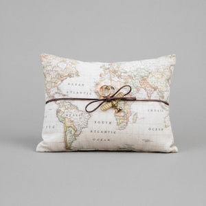 Può includere: Un cuscino quadrato con un design a mappa del mondo. Un cinturino in pelle marrone avvolge il cuscino, tenendo due anelli d'oro e un piccolo ciondolo a forma di aeroplano. Il cuscino è color crema con una stampa di mappa vintage.