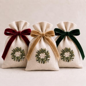 Può includere: Tre sacchetti in tessuto beige con coulisse, ciascuno caratterizzato da una ghirlanda ricamata verde con piccoli accenti bianchi. I sacchetti sono legati con nastri di velluto in rosso scuro, marrone dorato e verde bosco, formando un set.