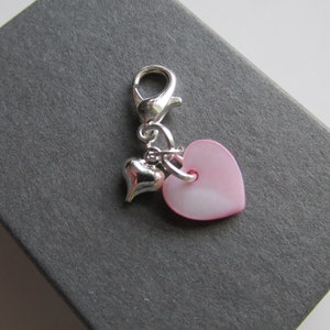 May include: A silver charm with a lobster clasp, featuring two heart-shaped pendants. One is a small, shiny silver heart, and the other is a larger, pale pink heart. The charm is likely for a bracelet or necklace.