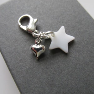 May include: A silver charm with a lobster clasp, a small heart pendant, and a white star pendant. The charm is designed for a bracelet or necklace, and is set against a grey background.