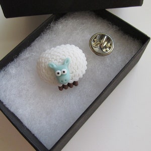 May include: A white sheep pin with blue eyes and a brown nose. The pin is in a black box with white stuffing.